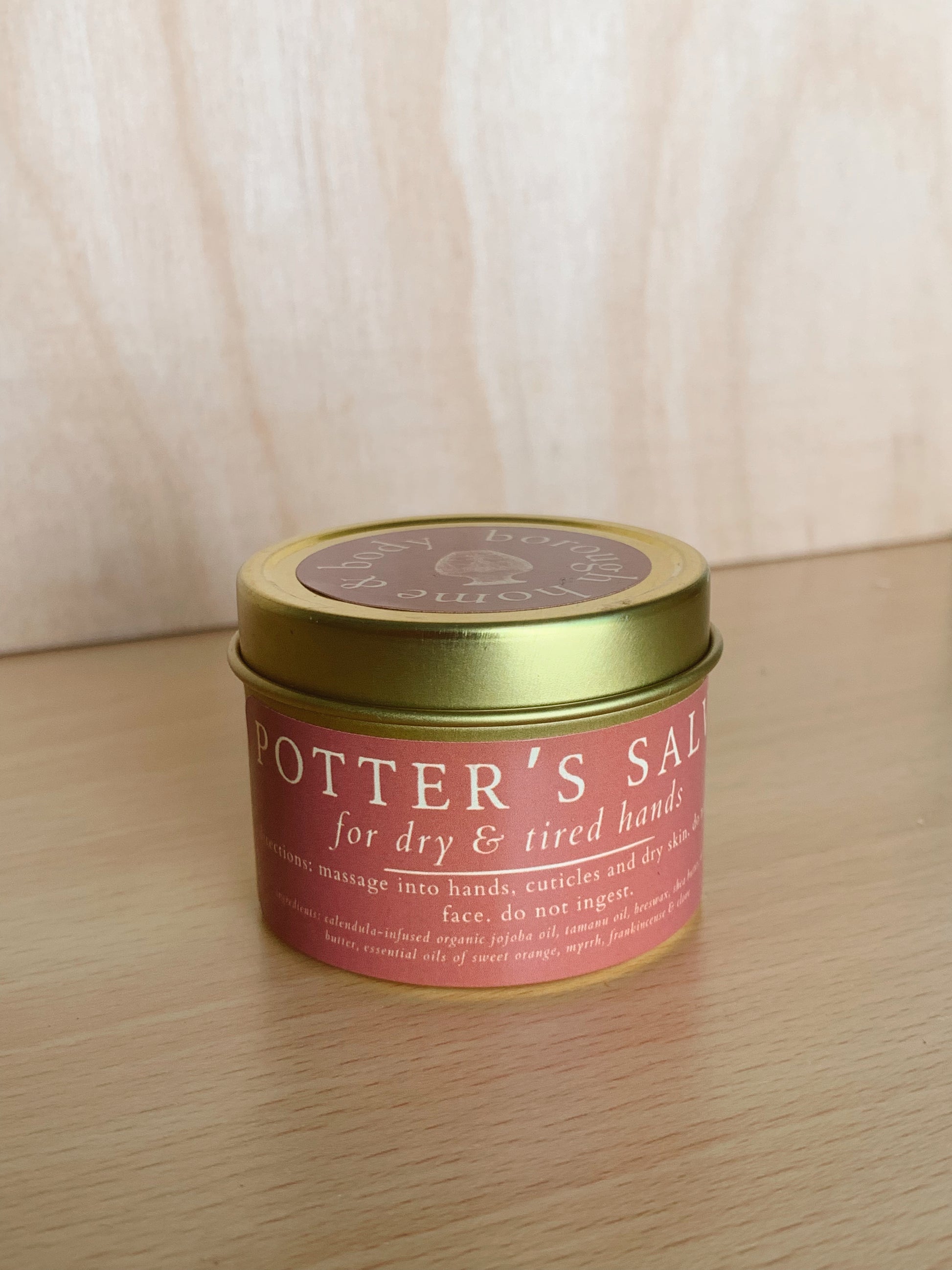 Tin of Potter's Sal for dry and tired hands on a wooden surface with a white curtain background.
