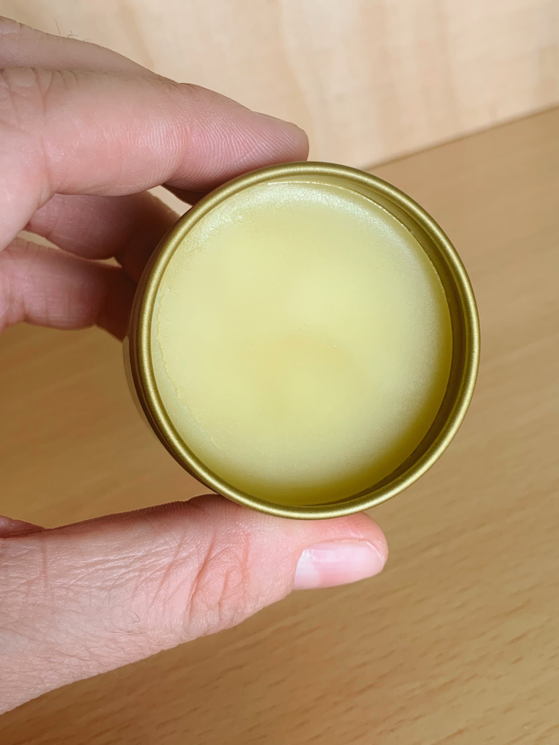 Hand holding a gold container with salve against a wooden surface