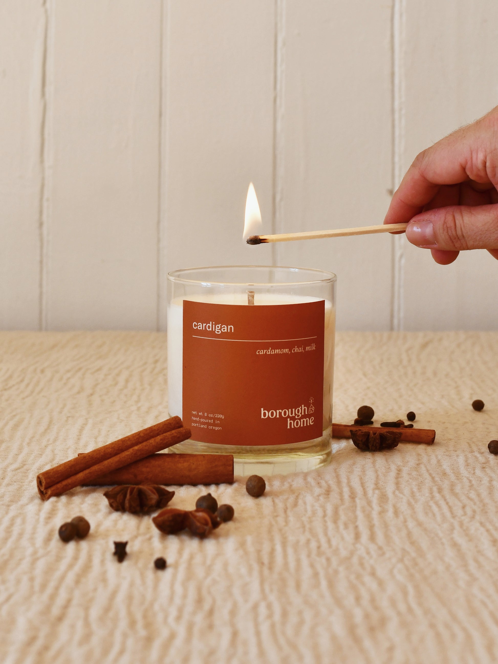 Cardigan candle with various warm spices and a hand holding a lit match, front view.